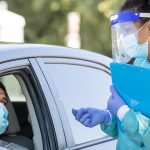 A nurse with full PPP stands outside a car with a clipboard and nasal swab, while a man with surgical mask sits in the driver's seat with the window down